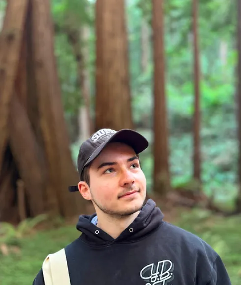 Young caucasian male wearing a black hoodie and cap, looking in the top right direction. You can see a forrest in the background.